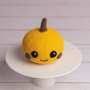 May include: A crocheted yellow pumpkin with a brown stem and a smiling face. The pumpkin has brown eyes and cheeks.