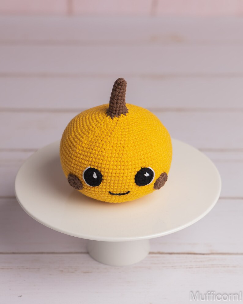 May include: A crocheted yellow pumpkin with a brown stem and a smiling face. The pumpkin has brown eyes and cheeks.