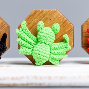 May include: A green crocheted bat with large wings, sitting on a wooden base. The bat is a handmade craft item.