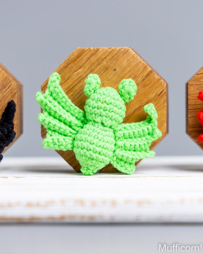 May include: A green crocheted bat with large wings, sitting on a wooden base. The bat is a handmade craft item.