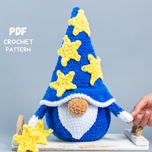 Könnte beinhalten: Ein handgefertigter blauer Häkelzwerg mit gelben Sternen auf Hut und Armen. Der Zwerg hat einen weißen Bart und eine braune Nase. Der Text "PDF CROCHET PATTERN" ist sichtbar. Der Zwerg steht auf einem weißen Tablett.