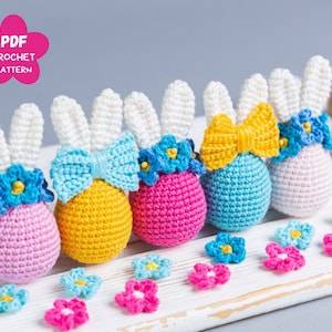 Peut inclure: Cinq décorations d'œufs de Pâques en forme de lapin crochetées en rose, jaune, bleu et blanc. Chaque lapin a un nœud et une couronne de fleurs. Les lapins sont disposés sur une surface en bois blanc.