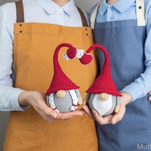 May include: Two crocheted gnomes with red hats and white beards holding hands. The gnomes are holding hands in a heart shape. The gnomes are in front of two people wearing aprons.