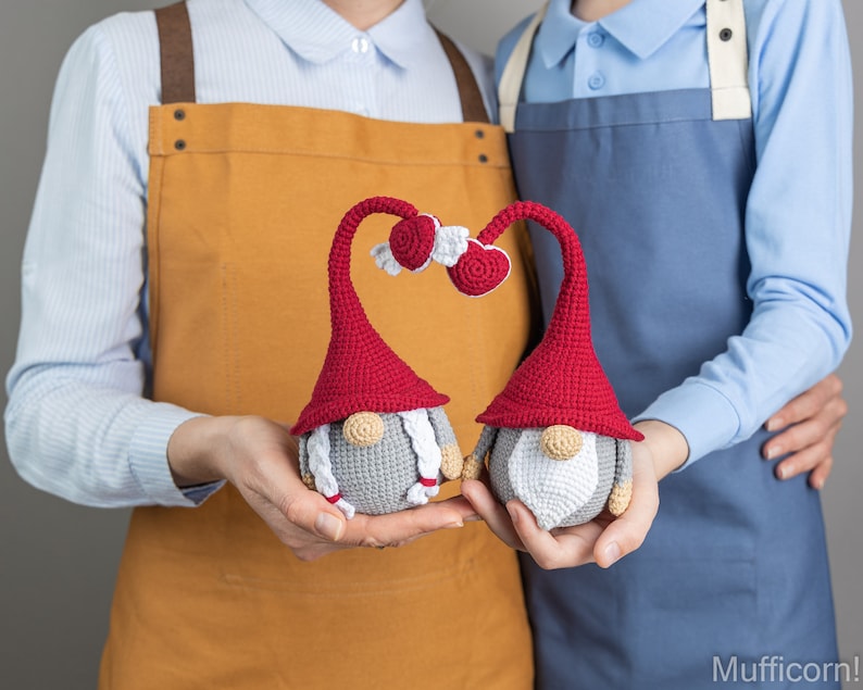 May include: Two crocheted gnomes with red hats and white beards holding hands. The gnomes are holding hands in a heart shape. The gnomes are in front of two people wearing aprons.
