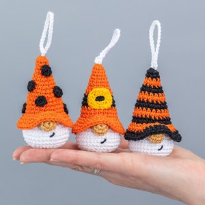 May include: Three handmade crochet gnome ornaments. Each gnome has a white body, a gold-colored nose, and a pointed hat in orange and black. One hat has black polka dots, another has a yellow buckle, and the third has black stripes.