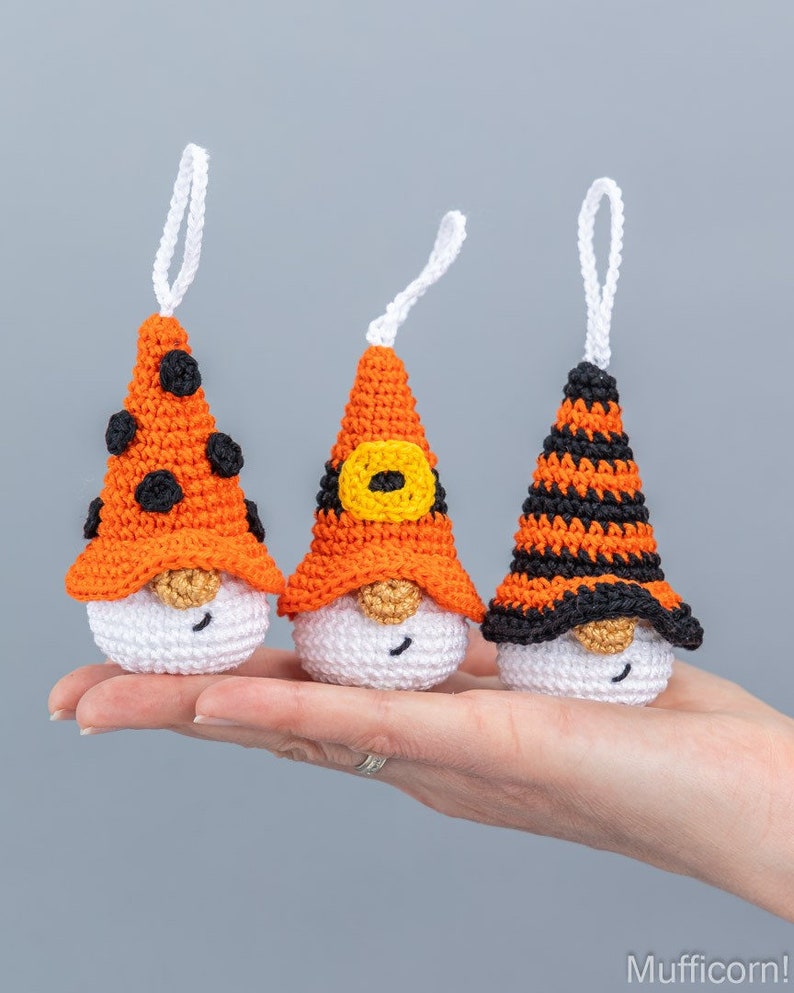 May include: Three handmade crochet gnome ornaments. Each gnome has a white body, a gold-colored nose, and a pointed hat in orange and black. One hat has black polka dots, another has a yellow buckle, and the third has black stripes.