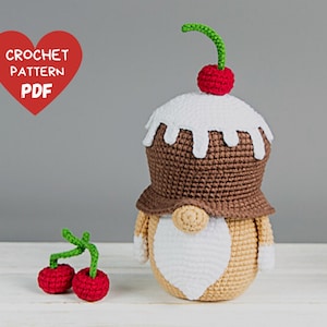 May include: Crochet pattern PDF for a brown and white gnome with a cherry on top. The gnome is wearing a brown hat with white icing and a white beard. There are two red cherries with green stems on a white surface.