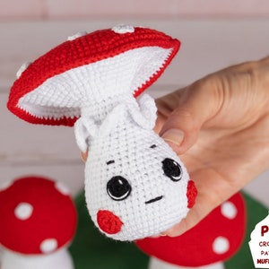May include: A crocheted mushroom amigurumi toy with a red cap and white body. The mushroom has a cute face with black eyes and a red nose. The toy is being held in a hand.