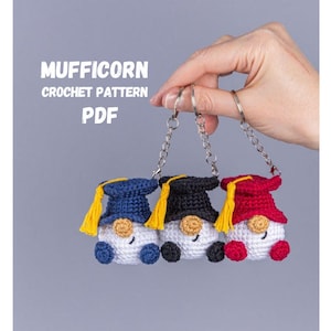May include: Three crocheted gnome keychains in blue, black, and red graduation caps with yellow tassels. The gnomes have white bodies, blue, black, and red caps, and are held by a silver keychain. The text "MUFFICORN CROCHET PATTERN PDF" is in the background.
