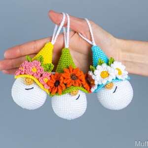 May include: Three crocheted gnome ornaments with different colored hats and flower crowns. The gnome on the left has a yellow hat with pink flowers, the middle gnome has a green hat with orange flowers, and the gnome on the right has a blue hat with white flowers.