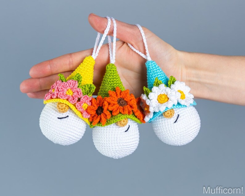May include: Three crocheted gnome ornaments with different colored hats and flower crowns. The gnome on the left has a yellow hat with pink flowers, the middle gnome has a green hat with orange flowers, and the gnome on the right has a blue hat with white flowers.