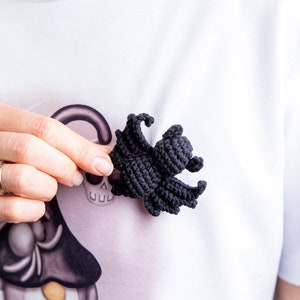 May include: A black crocheted spider with eight legs. The spider is being held by a hand in front of a white shirt with a Halloween-themed design.