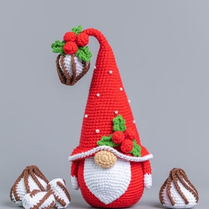 May include: A crocheted Christmas gnome wearing a red hat with white polka dots and a white beard. The gnome is holding a red and green holly sprig with red berries. There are three white and brown crocheted candy canes on the table.