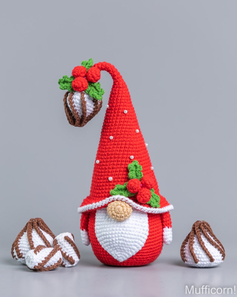 May include: A crocheted Christmas gnome wearing a red hat with white polka dots and a white beard. The gnome is holding a red and green holly sprig with red berries. There are three white and brown crocheted candy canes on the table.