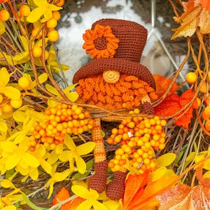 May include: A crocheted gnome wearing a brown hat with an orange flower on top. The gnome is surrounded by autumn foliage, including yellow flowers and orange leaves.