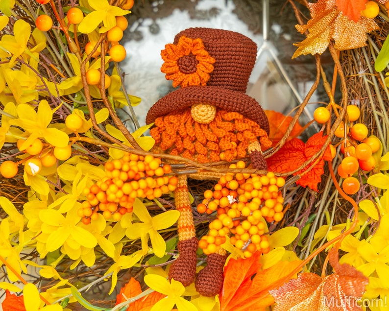 May include: A crocheted gnome wearing a brown hat with an orange flower on top. The gnome is surrounded by autumn foliage, including yellow flowers and orange leaves.