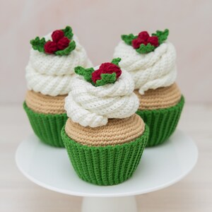 May include: Three crocheted cupcakes with green icing and red holly berries on top. The cupcakes are sitting on a white cake stand.