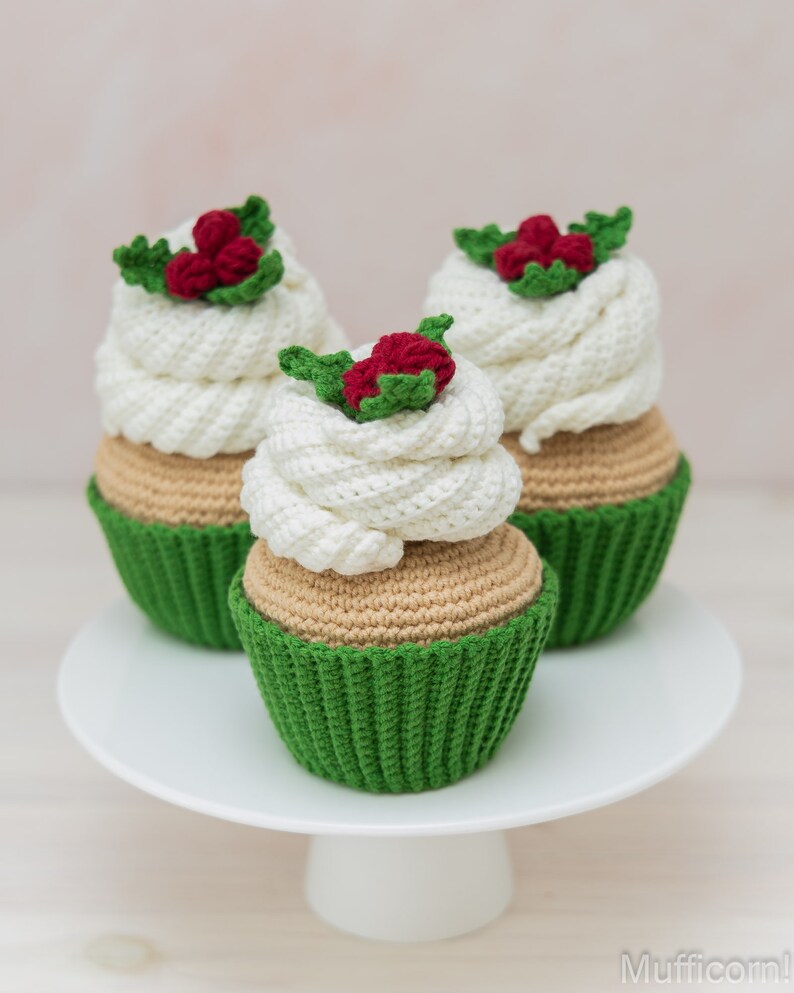 May include: Three crocheted cupcakes with green icing and red holly berries on top. The cupcakes are sitting on a white cake stand.