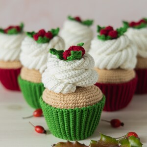 May include: A group of five crocheted cupcakes decorated with white frosting and red holly berries. The cupcakes are arranged in a row on a white surface with green holly leaves.