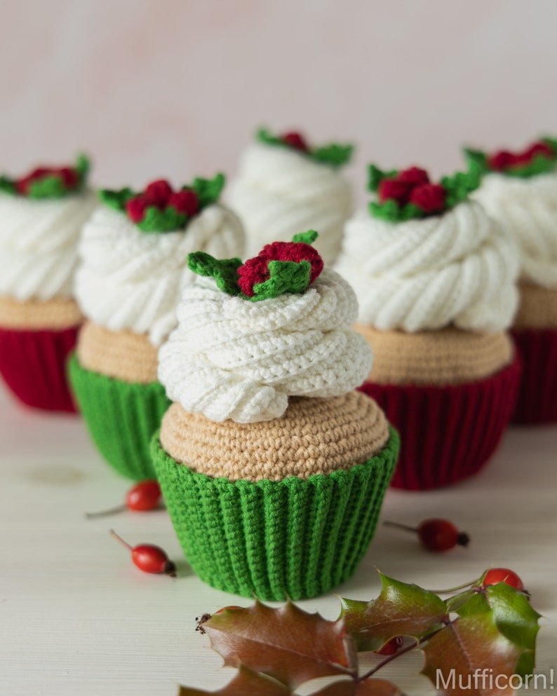 May include: A group of five crocheted cupcakes decorated with white frosting and red holly berries. The cupcakes are arranged in a row on a white surface with green holly leaves.