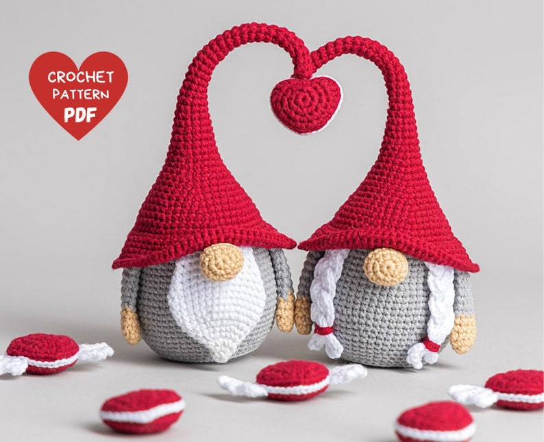 May include: Two crocheted gnomes in red and gray with white beards and braids. The gnomes are holding hands in the shape of a heart. The gnomes are surrounded by red and white candy canes.