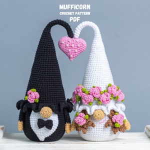 May include: A crocheted wedding gnome couple with a pink heart above them. The groom gnome is wearing a black suit with a white bow tie and the bride gnome is wearing a white dress with a pink flower crown.