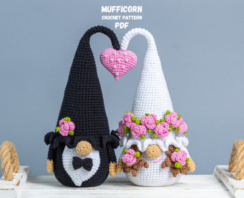May include: A crocheted wedding gnome couple with a pink heart above them. The groom gnome is wearing a black suit with a white bow tie and the bride gnome is wearing a white dress with a pink flower crown.