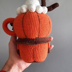 Puede incluir: Una taza de ganchillo con forma de calabaza con asa, detalles marrones y una cobertura de crema batida blanca. La taza naranja tiene un borde marr&oacute;n y un tallo marr&oacute;n. La taza est&aacute; hecha a mano y tiene un aspecto texturizado.