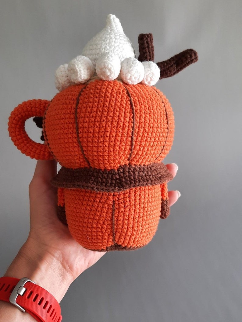 Puede incluir: Una taza de ganchillo con forma de calabaza con asa, detalles marrones y una cobertura de crema batida blanca. La taza naranja tiene un borde marr&oacute;n y un tallo marr&oacute;n. La taza est&aacute; hecha a mano y tiene un aspecto texturizado.