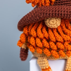 May include: A crocheted gnome with a brown hat and orange beard. The gnome is sitting on a white surface.