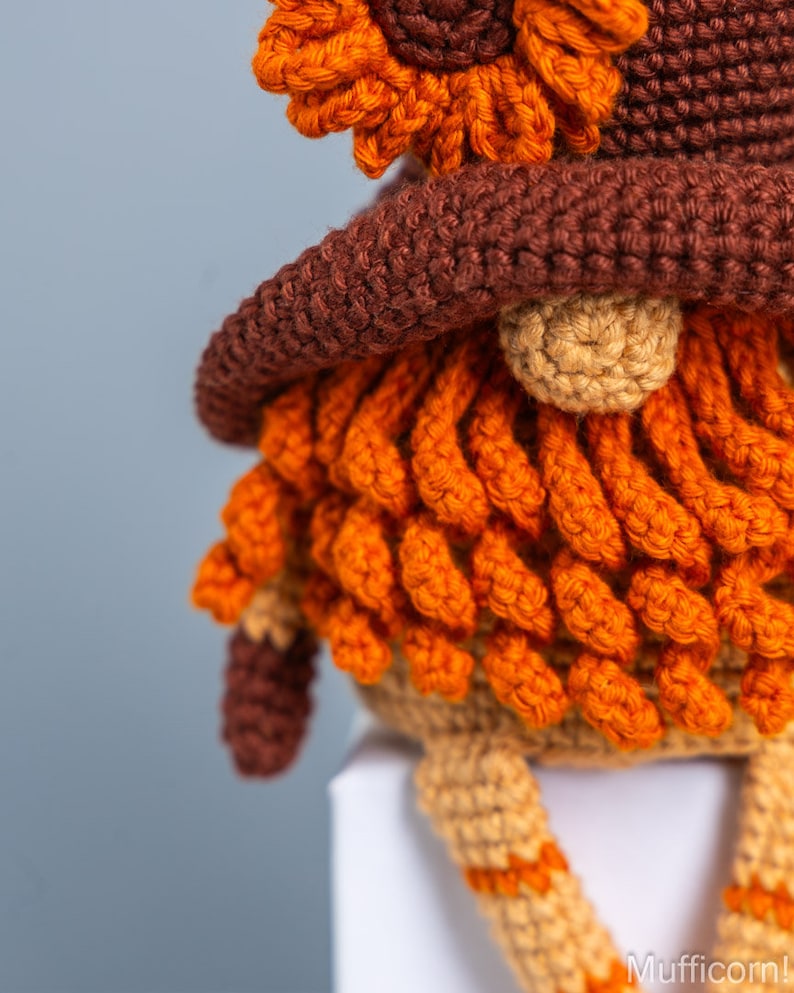 May include: A crocheted gnome with a brown hat and orange beard. The gnome is sitting on a white surface.