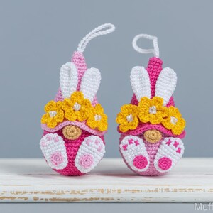 May include: Two pink and white crocheted gnomes with yellow flower crowns and white bunny ears. The gnomes are sitting on a white surface.