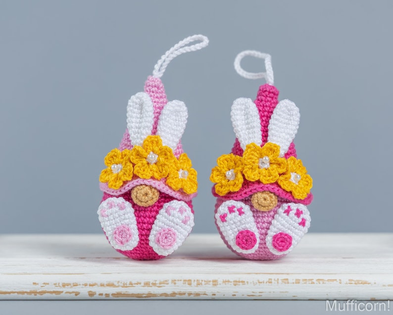 May include: Two pink and white crocheted gnomes with yellow flower crowns and white bunny ears. The gnomes are sitting on a white surface.