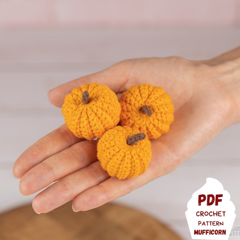 May include: Three crocheted orange pumpkins in a hand. The pumpkins are small and have a brown stem. The image includes the text "PDF Crochet Pattern Mufficorn".