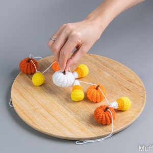 May include: A crocheted garland featuring orange and white pumpkins and candy corn. The garland is strung on a white cord and is hanging on a wooden surface.