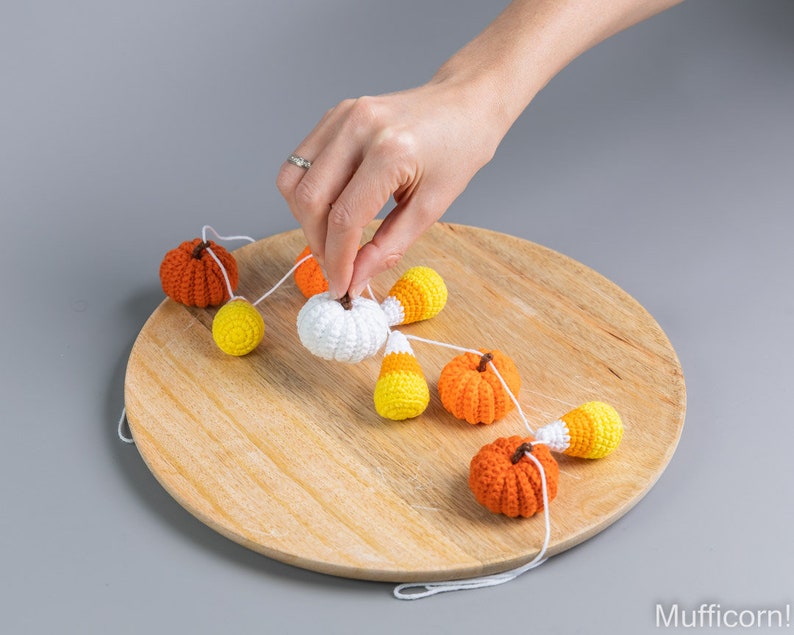May include: A crocheted garland featuring orange and white pumpkins and candy corn. The garland is strung on a white cord and is hanging on a wooden surface.