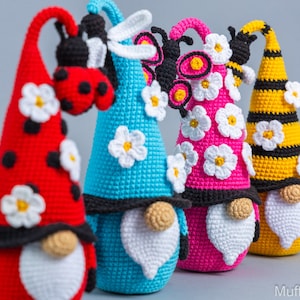 May include: Four crocheted gnomes with different insect themes. The gnomes are wearing hats with white daisy flowers and have white beards. The gnomes are red, blue, pink, and yellow with black accents.