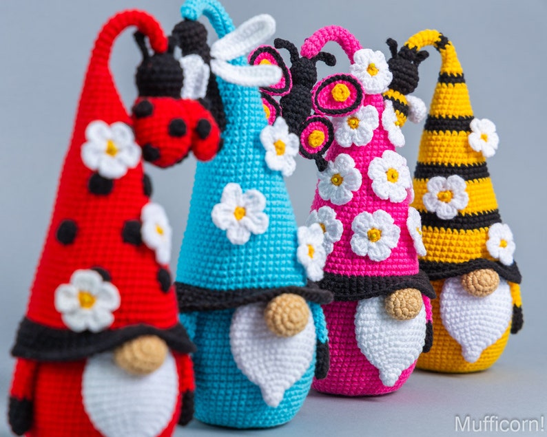 May include: Four crocheted gnomes with different insect themes. The gnomes are wearing hats with white daisy flowers and have white beards. The gnomes are red, blue, pink, and yellow with black accents.
