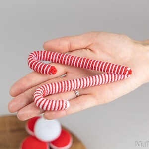 Puede incluir: Dos bastones de caramelo a rayas rojas y blancas se sostienen en una mano. Los bastones de caramelo est&aacute;n hechos de un material suave y tienen forma curva. Los bastones de caramelo son una decoraci&oacute;n festiva.