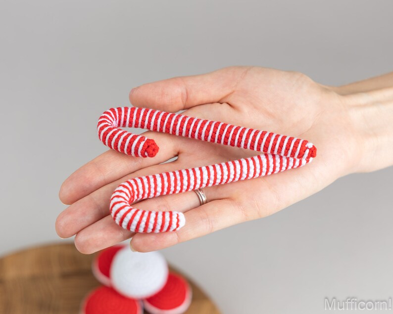 Puede incluir: Dos bastones de caramelo a rayas rojas y blancas se sostienen en una mano. Los bastones de caramelo est&aacute;n hechos de un material suave y tienen forma curva. Los bastones de caramelo son una decoraci&oacute;n festiva.