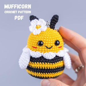 May include: A crocheted bee-shaped keychain with the text "MUFFICORN CROCHET PATTERN PDF". The bee has a yellow body with black stripes, black eyes, and a white flower on its head. It is held in a person's hand.