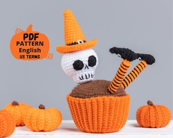 May include: Crocheted Halloween amigurumi toy of a skeleton wearing a witch's hat and sitting in a cupcake. The cupcake is orange and brown with black and orange striped legs sticking out. There are three small orange pumpkins in the background.