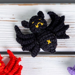 May include: A black crocheted bat with yellow eyes and a black cross on its chest. The bat is lying on a white wooden surface.