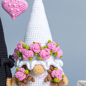 May include: A white crocheted gnome with a pink flower crown and pink flowers on its cheeks. The gnome is holding a pink heart with white pearl accents.