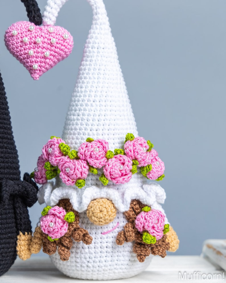 May include: A white crocheted gnome with a pink flower crown and pink flowers on its cheeks. The gnome is holding a pink heart with white pearl accents.