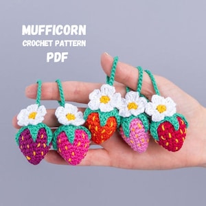 Könnte beinhalten: Fünf handgefertigte gehäkelte Erdbeer-Ornamente in Rot-, Rosa- und Lilatönen, jeweils mit einer weißen Blume und grünen Blättern. Der Text "MUFFICORN CROCHET PATTERN PDF" ist oben zu sehen.