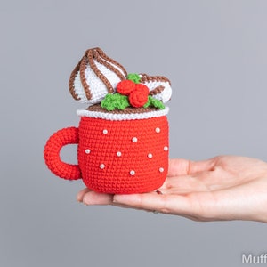 May include: A crocheted red mug with a white crocheted marshmallow and green holly on top. The mug is decorated with white beads.