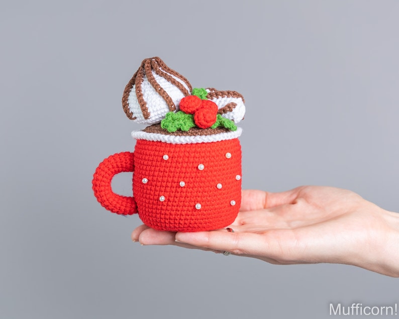May include: A crocheted red mug with a white crocheted marshmallow and green holly on top. The mug is decorated with white beads.