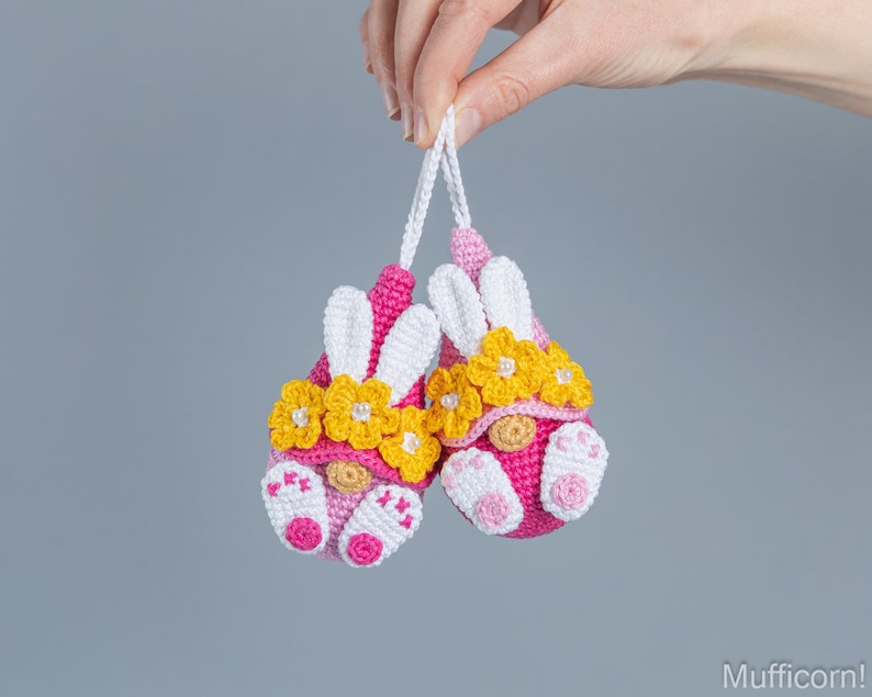 May include: Two pink crocheted gnomes with white bunny ears and yellow flower crowns. The gnomes are hanging from a string.