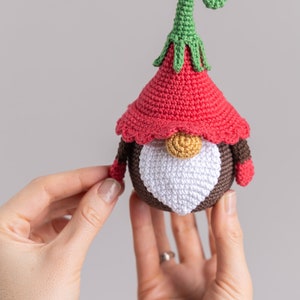 May include: A crocheted gnome ornament with a red hat, brown body, white beard, and a green curly hat.
